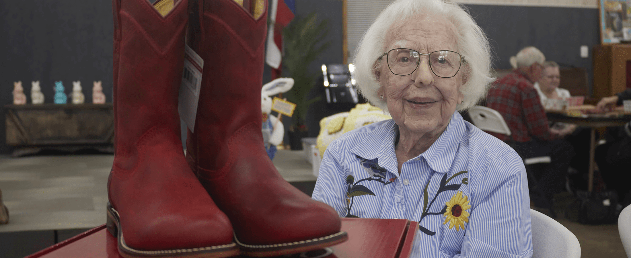 A lady with white hair, glasses, and a blue and white striped shirt with flowers posing and smiling next to her first pair of red cowboy roper boots. 